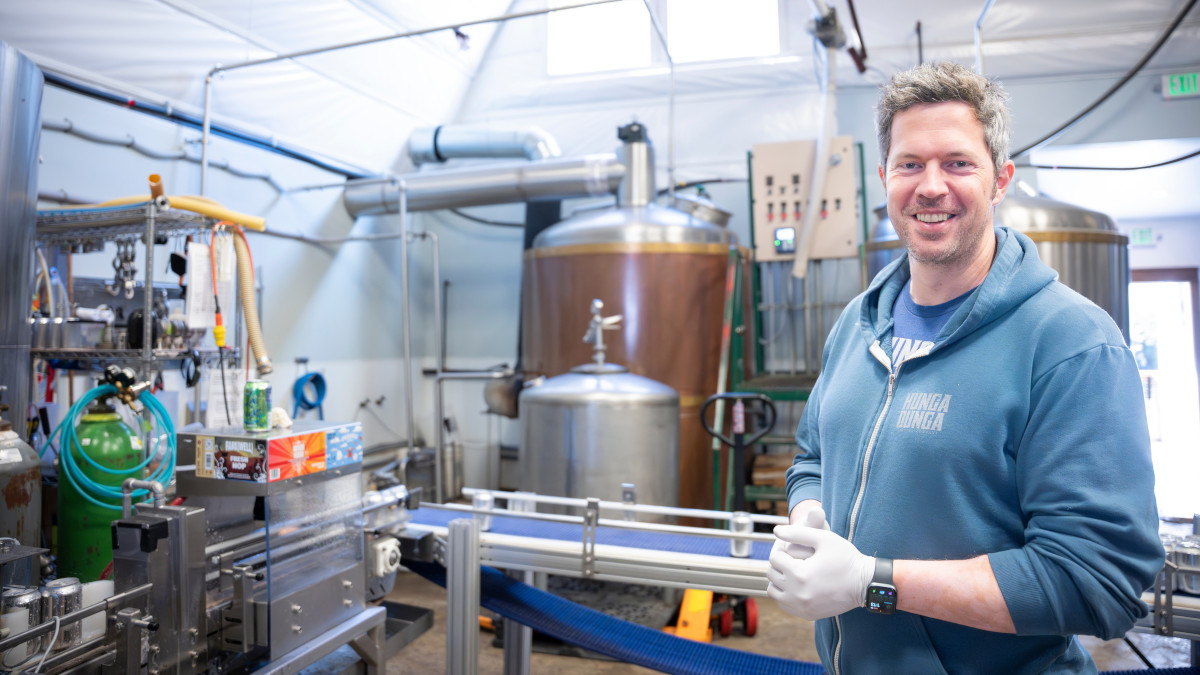 Man in blue sweatshirt stands in front of stainless steel equipment used to brew beer.